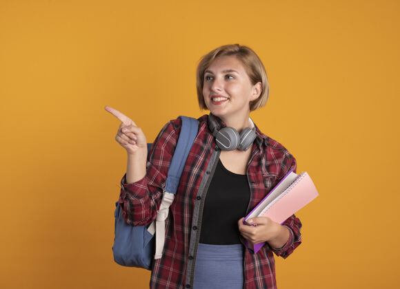 笔记本微笑着的年轻斯拉夫女学生戴着耳机 背着背包 拿着书和笔记本 看着并指着旁边壁板微笑背包