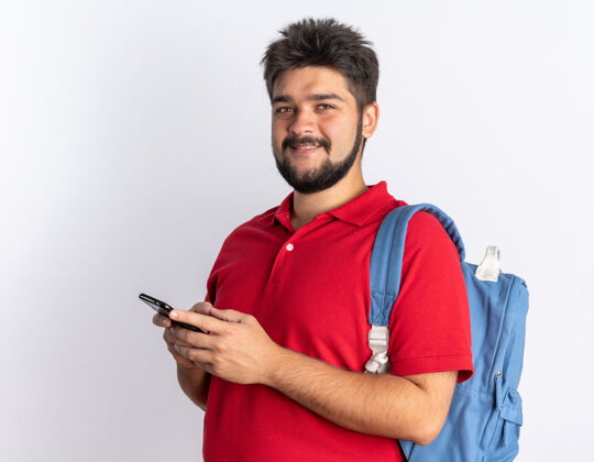 自信年轻的留着胡子的学生 穿着红色马球衫 背着背包 拿着智能手机 微笑着 自信 快乐 积极的站着微笑学生站着