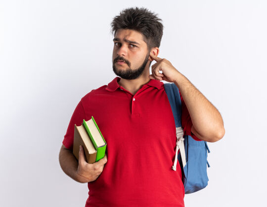 胡子年轻的留着胡子的学生 穿着红色马球衫 背着书包 拿着笔记本 站着严肃的脸 看起来很有趣红色学生阴谋