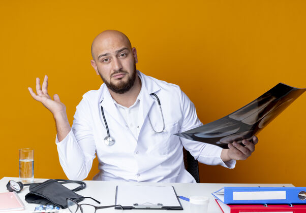 听诊器年轻的秃头男医生穿着医用长袍和听诊器坐在办公桌旁给人留下深刻印象印象X光办公桌