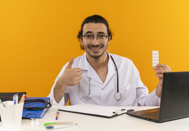 眼镜面带微笑的年轻男医生 戴着医用眼镜 穿着医用长袍 听诊器坐在办公桌旁医疗持有复制