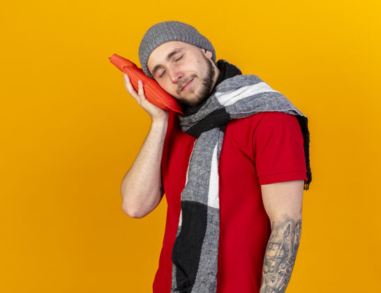 水高兴的年轻白人病人戴着冬天的帽子和围巾把头放在橙色的热水瓶上围巾年轻热