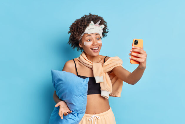 醒着积极的卷发年轻的非裔美国妇女通过智能手机自拍微笑愉快地将胶原蛋白贴在眼睛下面穿着睡衣抱枕隔离在蓝色的工作室墙上室内短信睡衣