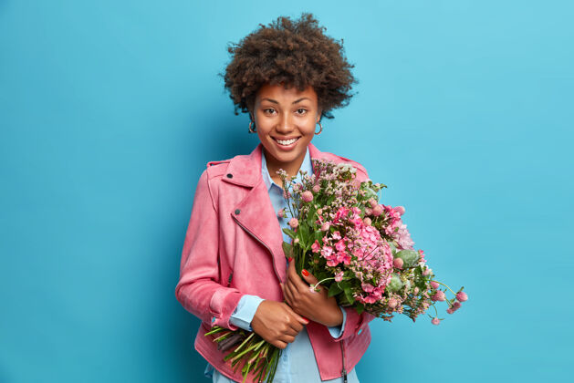 芳香快乐微笑的美国黑人年轻女子手持一大束鲜花横拍庆幸春天终于来了穿着时尚的粉色夹克隔着蓝色的墙壁开花卷曲事件