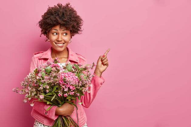 节日积极的年轻卷发女子摆出漂亮的花束指向空白显示广告内容穿着夹克隔离在粉红色的墙壁开花天咬