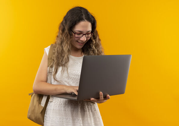 表情面带微笑的年轻漂亮的女学生戴着眼镜 背着包用笔记本电脑隔离在黄色的墙上漂亮市民眼镜