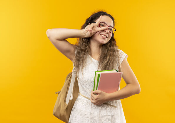 黄微笑着的年轻漂亮的女学生戴着眼镜 背着书包 拿着笔记本 做着隔离在黄色墙上的和平标志抱人年轻