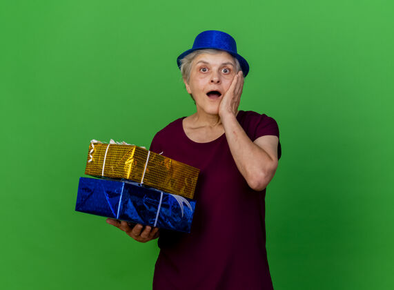 女人惊讶的老妇人戴着聚会帽把手放在脸上拿着绿色的礼盒聚会盒子老人