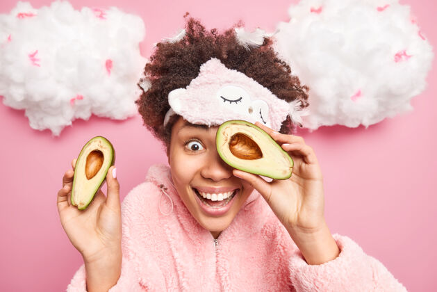 微笑正面的非裔美国女性的水平镜头使用天然的cosmetc产品 用鳄梨覆盖脸部 半个微笑 穿着睡衣 隔离在粉红色的墙上护理人皮肤