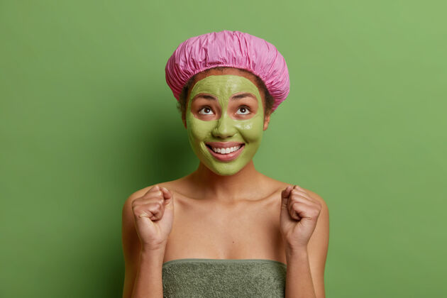 治疗积极的女人高兴地看着上方 握紧拳头等待特殊的东西戴着浴帽和毛巾在身体周围涂上滋养鳄梨面膜隔离在绿色的墙上拳头健康模特