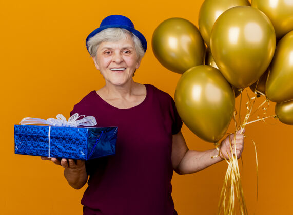 气球高兴的老妇人戴着党的帽子举行氦气球和礼品盒上的橙色派对氦气请穿