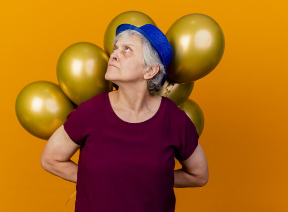 女人戴着派对帽的恼怒的老妇人在身后举着氦气球仰望橙色帽子穿气球