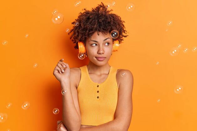 明亮沉思的年轻美国黑人妇女专注在一边 通过立体声耳机听音乐 穿着休闲服 表情深邃 喜欢在橙色的墙上唱着歌词美罗曼表情音乐
