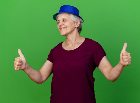 拇指戴派对帽的老太太高兴地竖起大拇指 两只手放在绿色上女人帽子老人