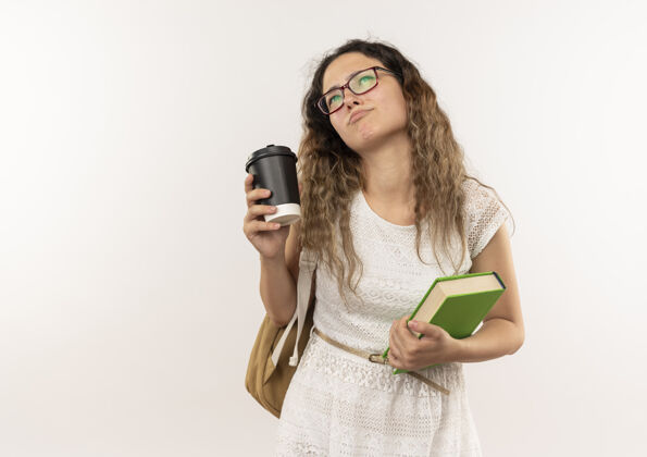 脸体贴的年轻漂亮的女学生戴着眼镜 背着书包 抬起头 手里拿着咖啡杯和书 孤零零地站在白色的墙上思想市民抱
