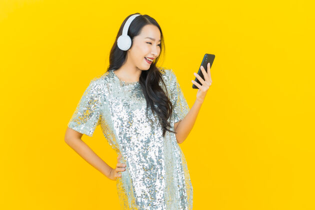 女士肖像美丽的亚洲年轻女子与耳机和智能手机听音乐的黄色放松可爱享受