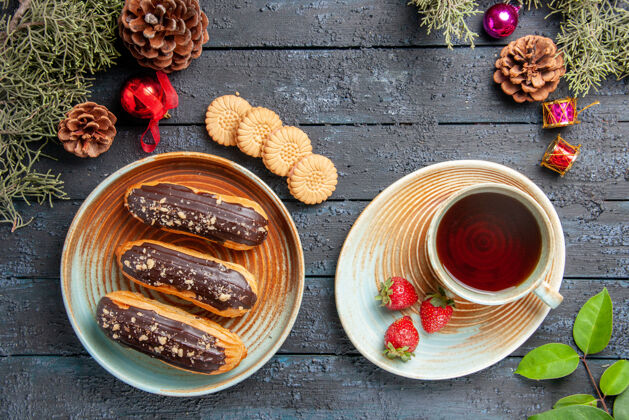 茶俯瞰图：椭圆形盘子上放着巧克力 茶托上放着一杯茶和草莓 松果 圣诞玩具 冷杉树 黑色木质地面上放着饼干黑暗玩具木头