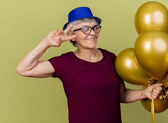 胜利戴着聚会帽 戴着眼镜 手持氦气球的老妇人高兴地站在那里 示意胜利请标志聚会