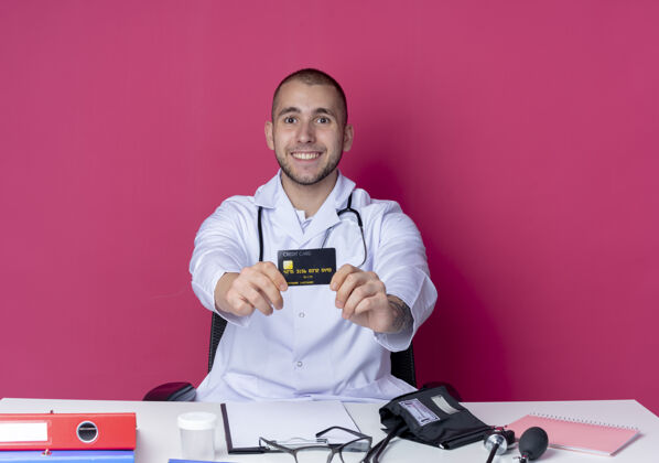 工作面带微笑的年轻男性医生 穿着医用长袍 听诊器 坐在办公桌旁 工作工具伸向隔离在粉色墙壁上的前方 信用卡伸向前方年轻人桌子人