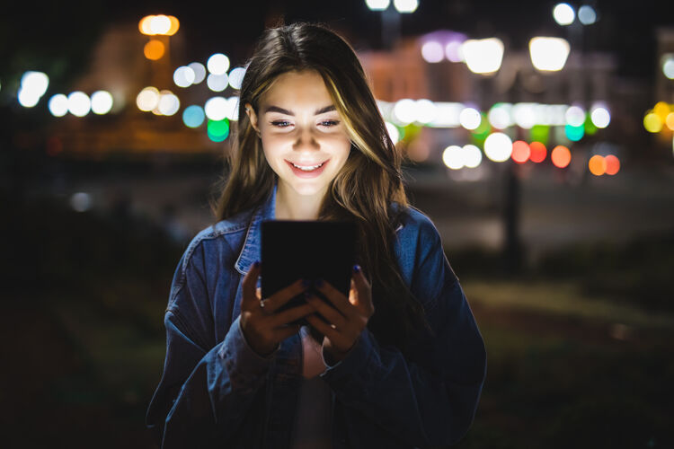 年轻美丽的女人侧视图使用数字平板电脑在城市街道上的夜晚时尚女孩使用