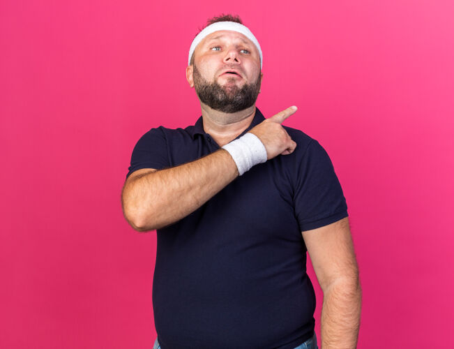 穿悲伤的成年斯拉夫运动型男子戴着头带和腕带 指向粉色墙壁上隔离的一侧 并留有复制空间运动男人成人