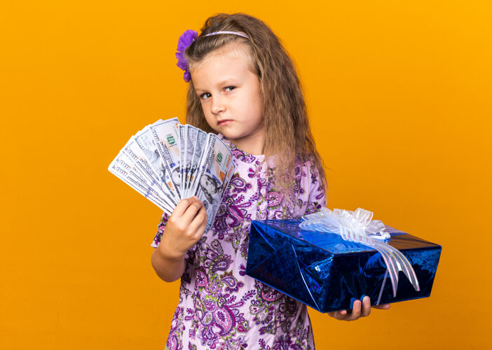 自信自信的金发小女孩拿着礼品盒和钱孤立在橙色墙上的复制空间金发孤立女孩