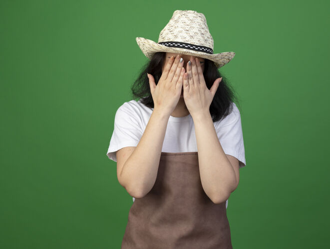 黑发悲伤的年轻黑发女园丁戴着眼镜和制服戴着园艺帽把手放在隔离在绿色墙上的脸上姿势人帽子