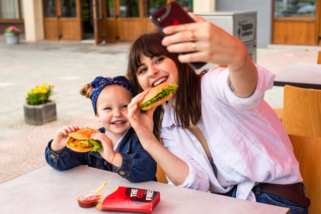 快餐年轻妈妈带着小女孩吃汉堡包在街边咖啡馆自拍美丽可爱孩子