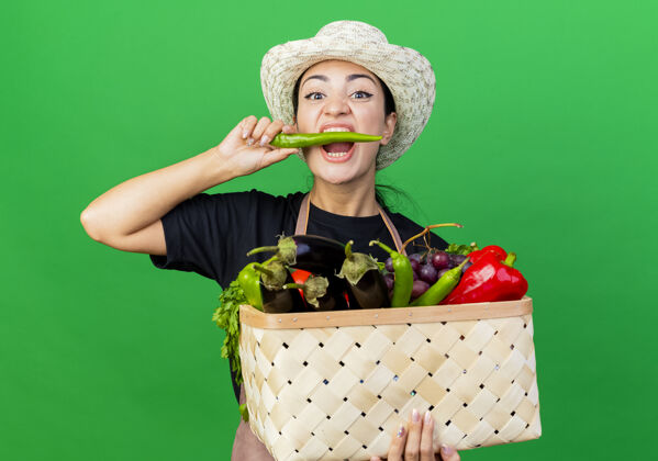 人年轻漂亮的女园丁围着围裙 戴着帽子 手里拿着装满蔬菜的篮子 咬着青椒站在绿色的墙上咬辣椒花园