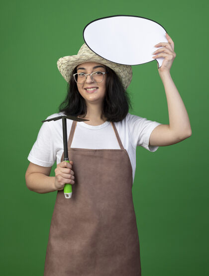 锄头微笑着的年轻黑发女园丁戴着眼镜和制服 戴着园艺帽 拿着锄头耙和隔离在绿色墙上的讲话泡泡人年轻眼镜