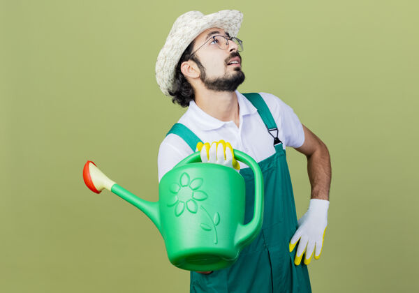 脸年轻的留着胡须的园丁 穿着连体衣 戴着帽子 手里拿着水罐子 站在浅绿色的墙上 困惑地抬头望着年轻人人表情