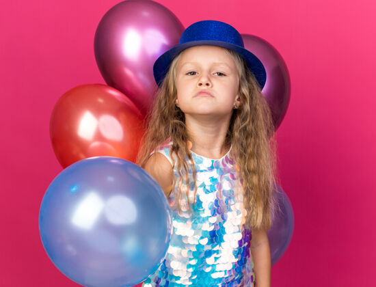 恼火恼怒的金发小女孩 戴着蓝色的派对帽 站在粉色墙上的氦气球上 还有复制空间女孩帽子站