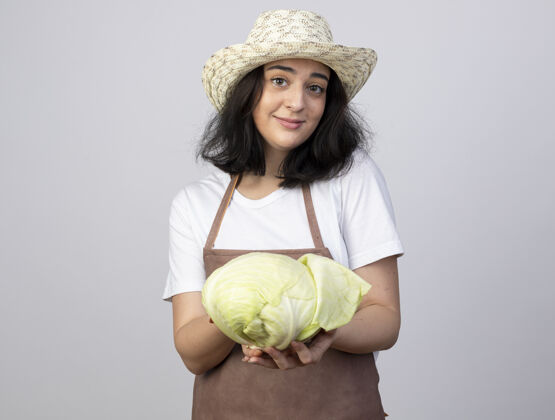 卷心菜年轻的黑发女园丁穿着制服 戴着园艺帽 抱着隔离在白墙上的卷心菜花园女年轻