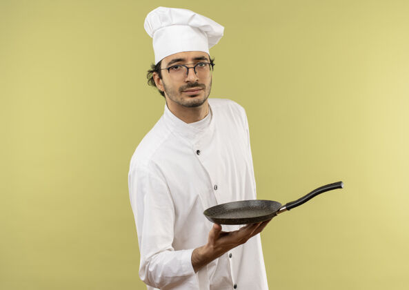 男性自信的年轻男厨师穿着厨师制服 戴着眼镜 拿着绿色背景的煎锅烹饪持有信心