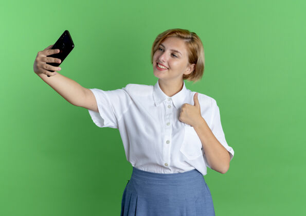 电话年轻微笑的金发俄罗斯女孩竖起大拇指看着手机向上女孩拇指
