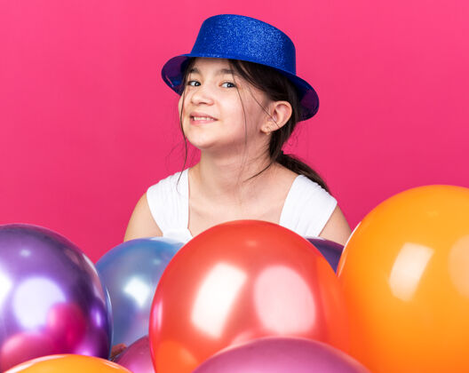 白种人微笑着的年轻白人女孩戴着蓝色的聚会帽站在粉红色的墙上 氦气球与复制空间隔离氦年轻帽子
