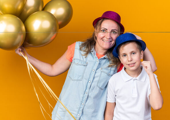 请高兴的年轻斯拉夫男孩戴着蓝色的派对帽 举起拳头 站在他母亲的身边 戴着紫色的派对帽 手里拿着氦气球 孤立地放在橙色的墙上 留着复制空间气球斯拉夫人站着