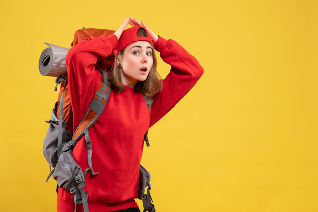 加入正面图：年轻的旅行妇女 背着红色的背包 双手举过头顶站在黄色的墙上头手帽子