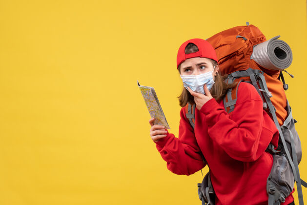 成人正面图：年轻的女性徒步旅行者 背着背包 戴着面具 手放在下巴上拿着地图背包前面地图