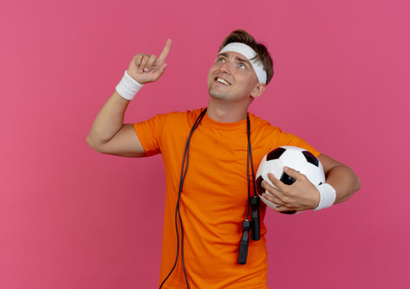 人面带微笑的年轻英俊的运动型男子戴着头带和腕带 脖子上系着跳绳 手里拿着足球 孤零零地指着粉色的墙上英俊头带周围