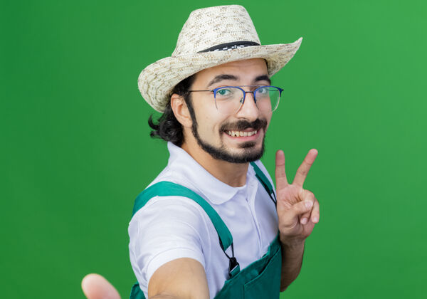 微笑年轻的留着胡须的园丁 穿着连体衣 戴着帽子 微笑着在绿色的墙壁上展示v形标志市民人Vsign