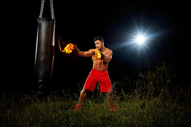 攻击运动员是在黑色的夜晚训练背景下 肌肉发达的拳击手套在夜间训练中 在户外训练 在草地上打拳击 从右手的黑色拳击袋里踢出来工作运动员激烈