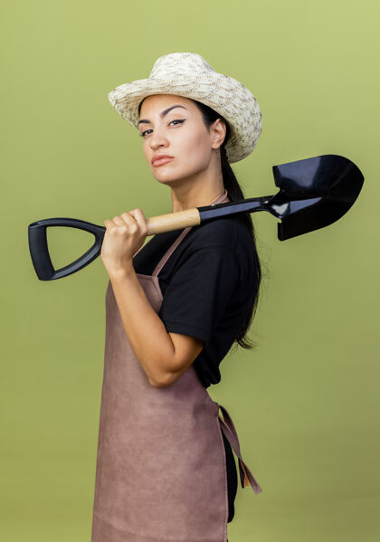 站着年轻漂亮的女园丁 围着围裙 戴着帽子 拿着铲子 严肃地看着前面 站在淡绿色的墙上帽子姿势铲子