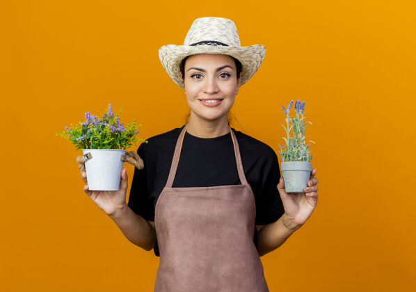 人年轻漂亮的女园丁 围着围裙 戴着帽子 手里拿着盆栽植物 站在橘色的墙上 面带微笑地看着前面围裙衣服女人