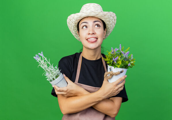 舌头年轻漂亮的女园丁 围着围裙 戴着帽子 手里拿着盆栽植物 站在绿色的墙壁上 仰望伸出的舌头人帽子脸