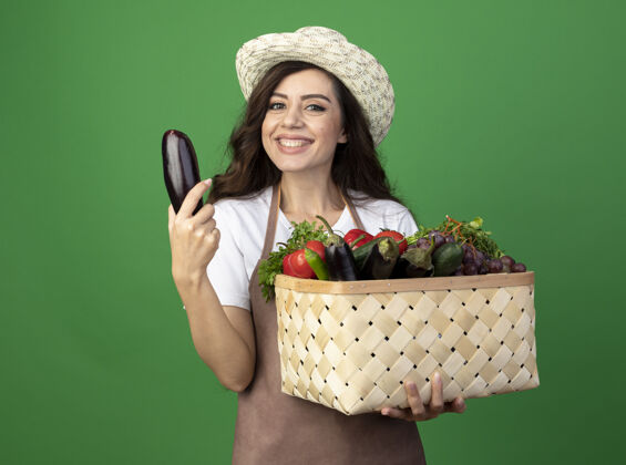 茄子身着制服 头戴园艺帽 面带微笑的年轻女园丁把菜篮子和茄子隔离在绿色的墙上园艺人年轻人