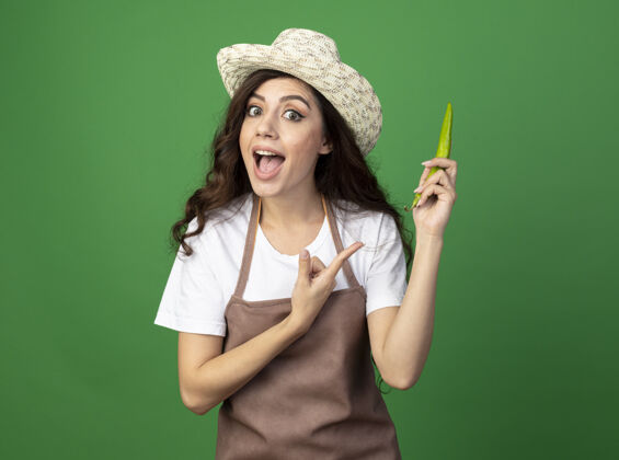 人穿着制服的年轻女园丁戴着园艺帽 指着隔离在绿墙上的辣椒印象胡椒女