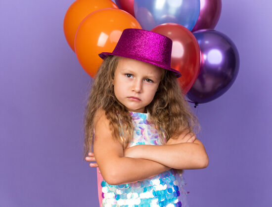 站自信的金发小女孩 戴着紫色派对帽 双臂交叉站在氦气球前 氦气球被隔离在紫色的墙上 留着复制空间前面金发女孩