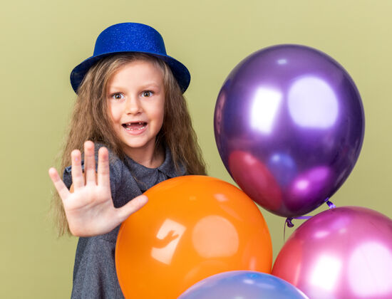 停惊讶的金发小女孩戴着蓝色派对帽站在那里 手里拿着氦气球 在橄榄绿的墙上做着停车标志的手势橄榄气球站