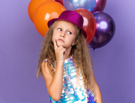 小困惑的金发小女孩 戴着紫色的派对帽 手指放在下巴上 看着站在紫色墙上的氦气球前面的一边孤立手指氦